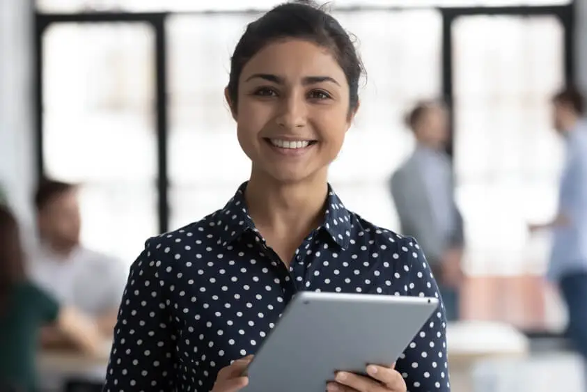 A smiling woman holding a tablet. She is standing in a modern office environment., appearing confident and happy, as if she has just solved a problem.